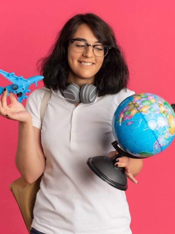 young-pleased-pretty-caucasian-schoolgirl-with-headphones-neck-wearing-glasses-back-bag-holds-toy-plane-globe-pink-with-copy-space